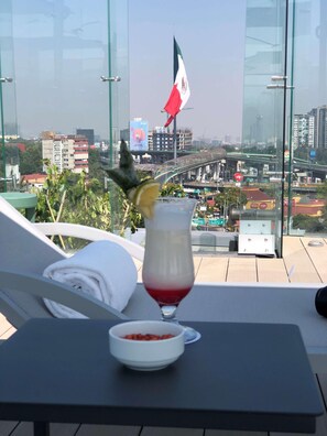 A glass of red drink with a lemon slice on a table, a bowl of snacks, and a view of a cityscape with buildings and a flag. - Galeria Plaza San Jeronimo (Mexico City)