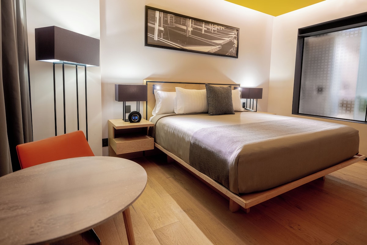 A modern hotel room with a bed, bedside tables, a desk, and a chair.