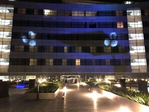 Un bâtiment moderne à la façade en verre, illuminé par un éclairage intérieur et des jeux de lumière décoratifs bleus.