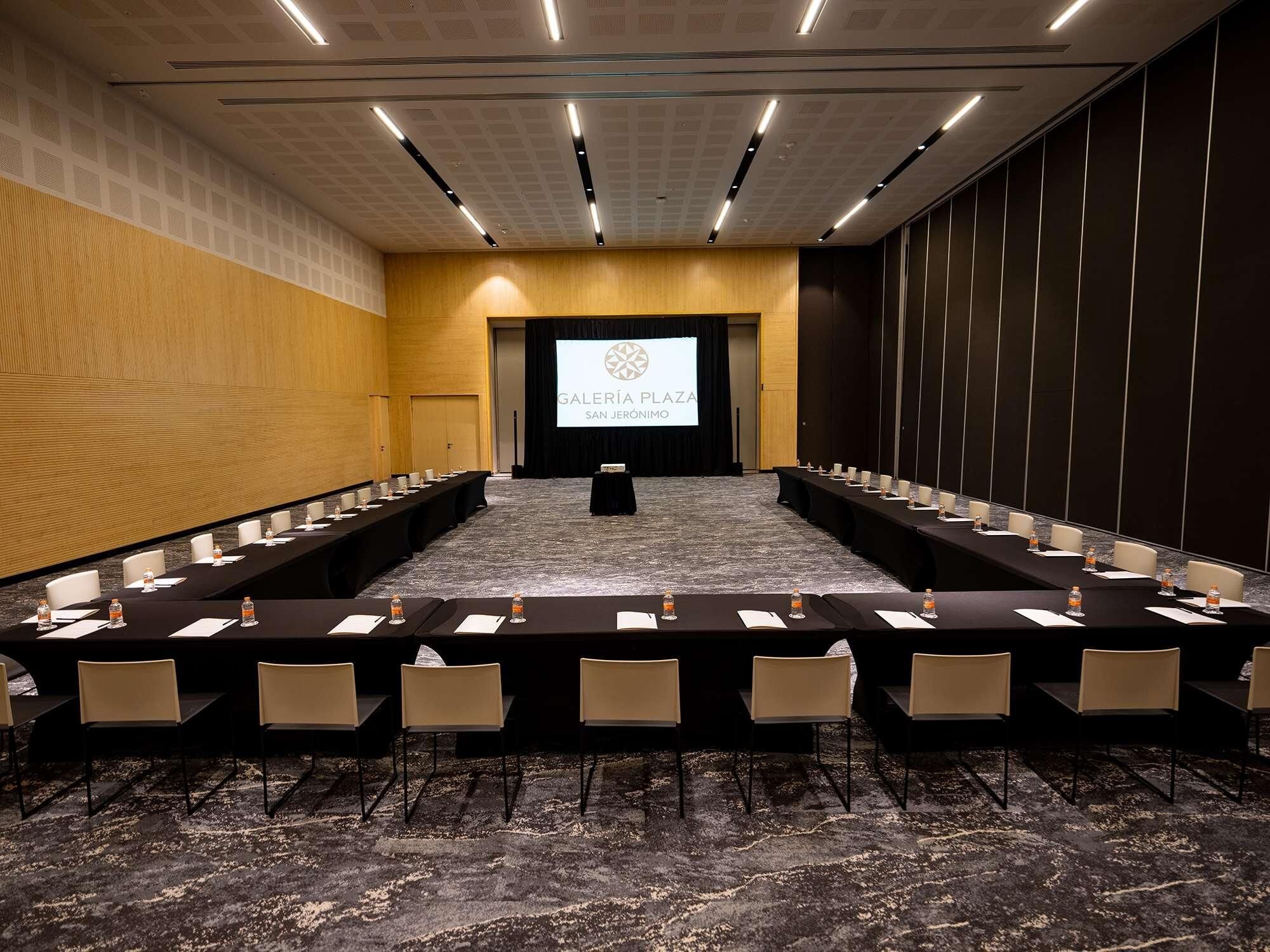 Un salón de conferencias con una larga mesa y sillas, una pantalla de proyector y piso alfombrado.