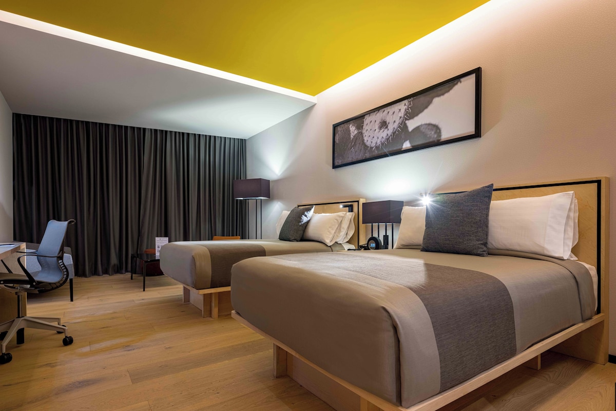 A hotel room with two beds, a desk, a chair, and a large window with curtains.