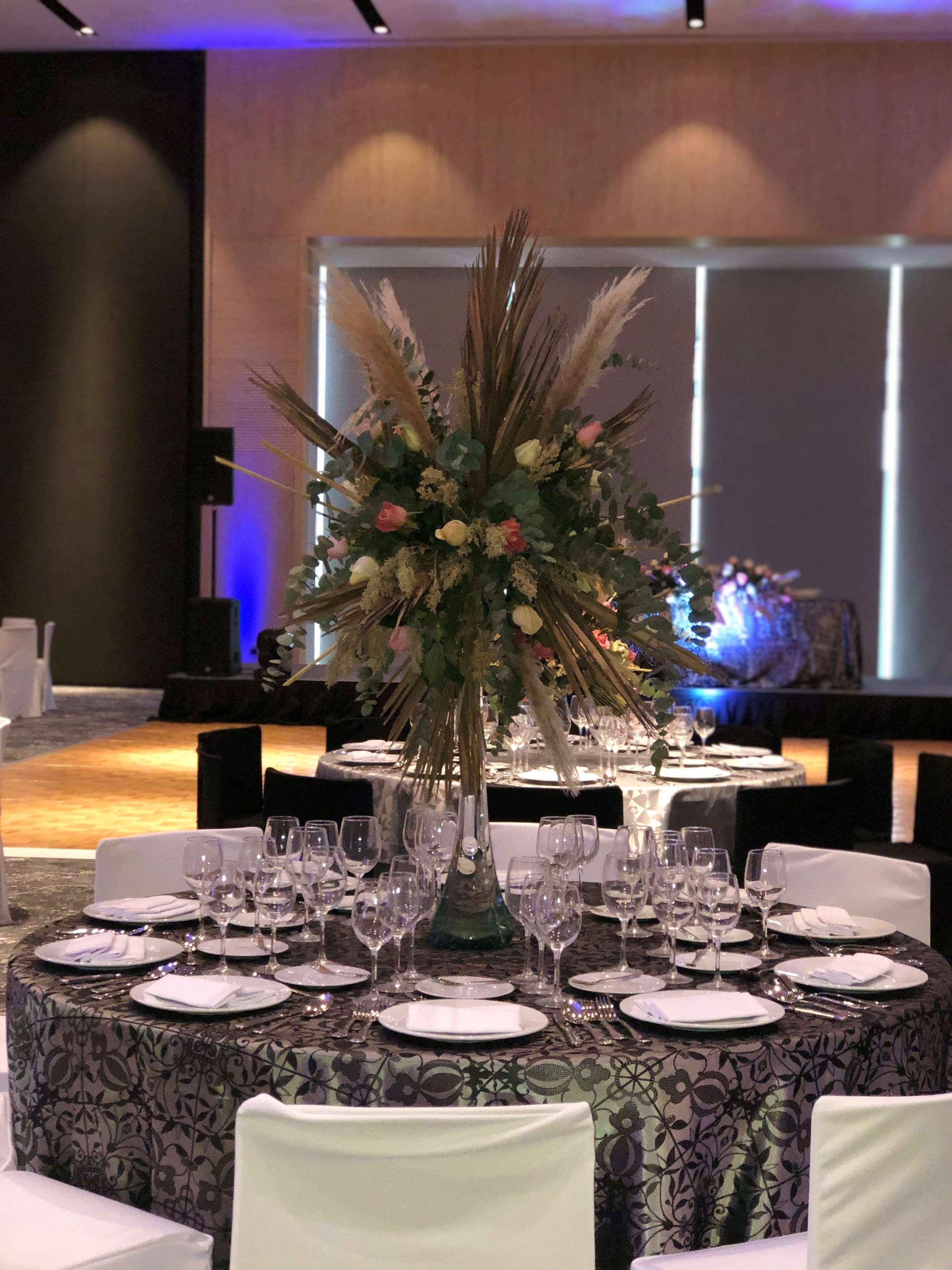 A banquet table set with white plates, wine glasses, and a centerpiece of flowers and pampas grass.