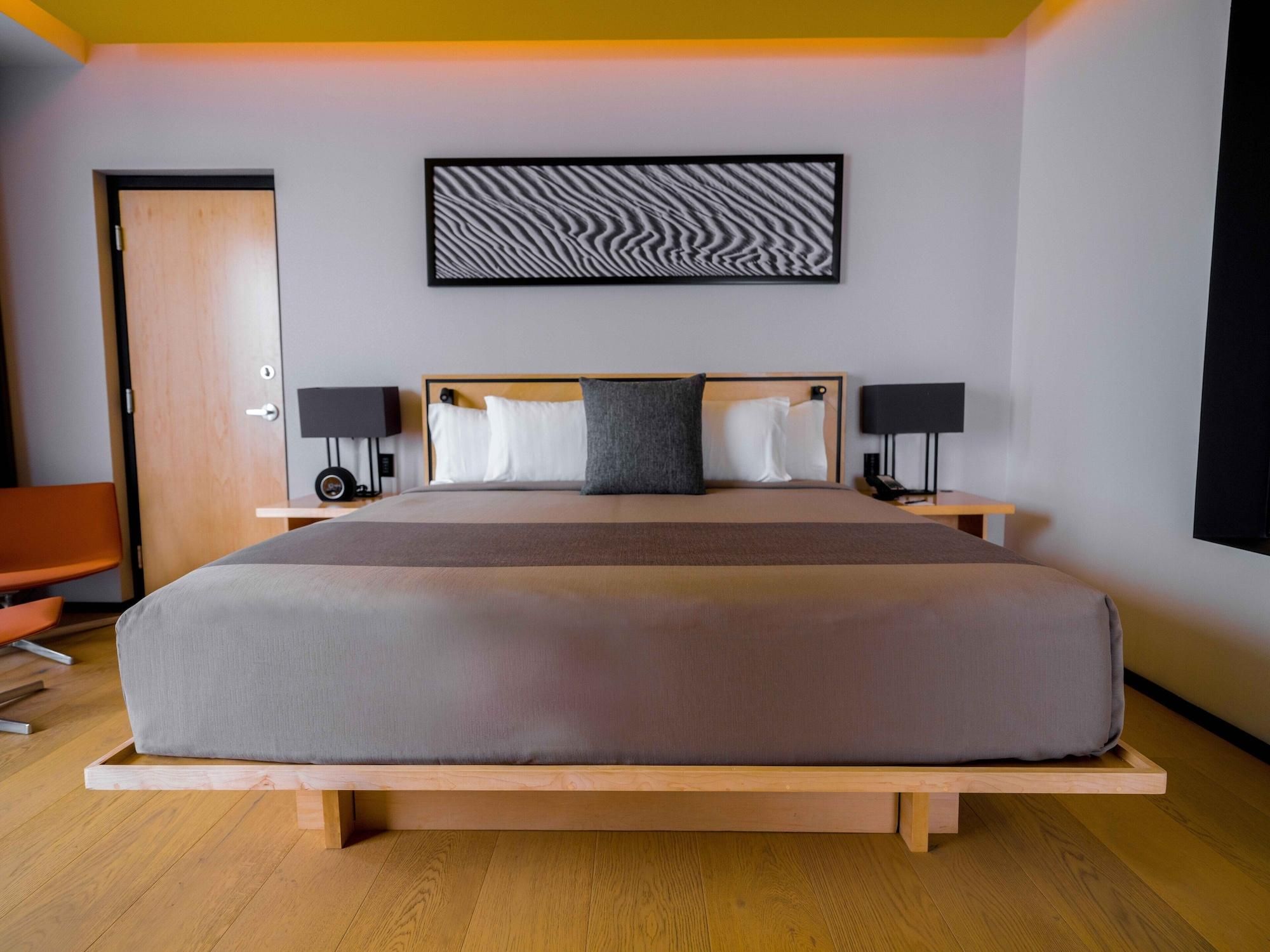 Habitación de hotel moderna con una cama grande, mesitas de noche con lámparas, un banco de madera, una puerta y una obra de arte en la pared.