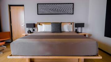 Habitación de hotel moderna con una cama grande, mesitas de noche con lámparas, un banco de madera, una puerta y una obra de arte en la pared.
