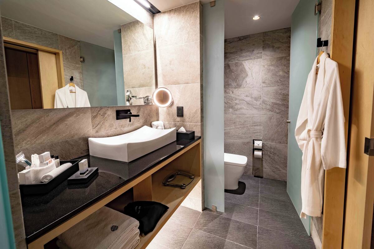 Une salle de bain moderne avec un lavabo aux lignes courbes, un miroir avec éclairage intégré, des toilettes et une robe suspendue à une porte.