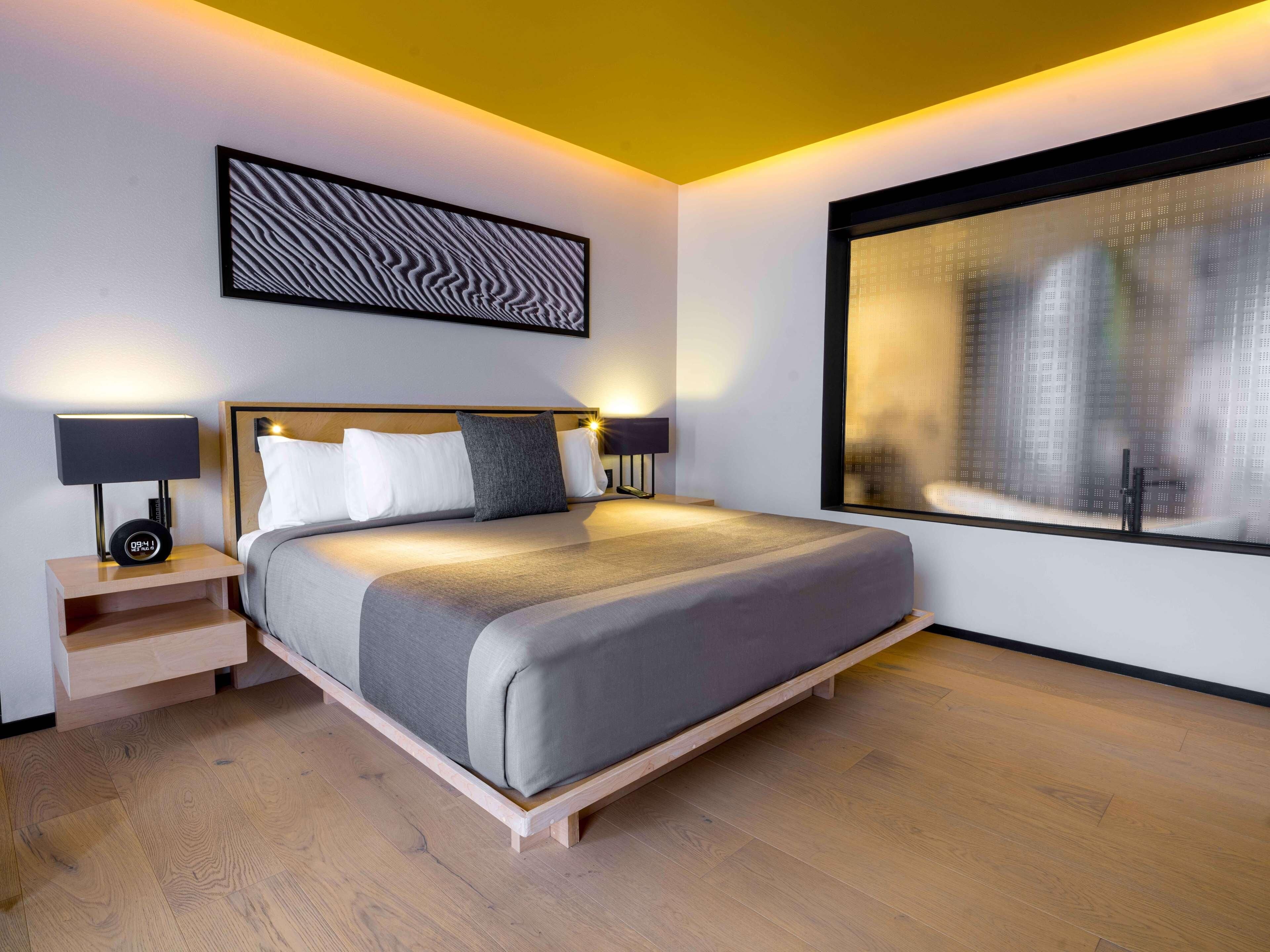 Una habitación de hotel moderna con una cama grande, mesitas de noche con lámparas, vista a la ciudad a través de una ventana y una obra de arte en la pared.