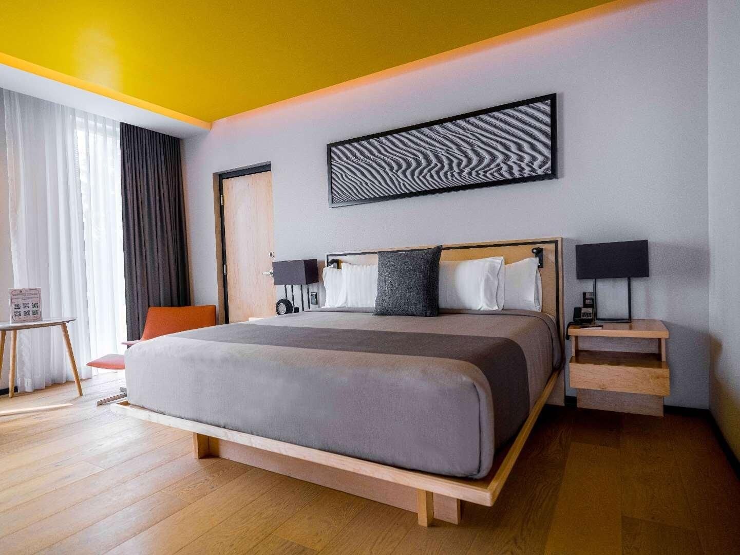 Une chambre d’hôtel moderne avec un grand lit, une tête de lit en bois, deux tables de chevet avec des lampes, un petit bureau et une chaise.