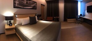A hotel room with a large bed, a desk, a chair, a TV, and a lamp.