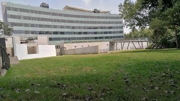 A modern building with a glass facade, a grassy area in front, and a pedestrian bridge in the background.