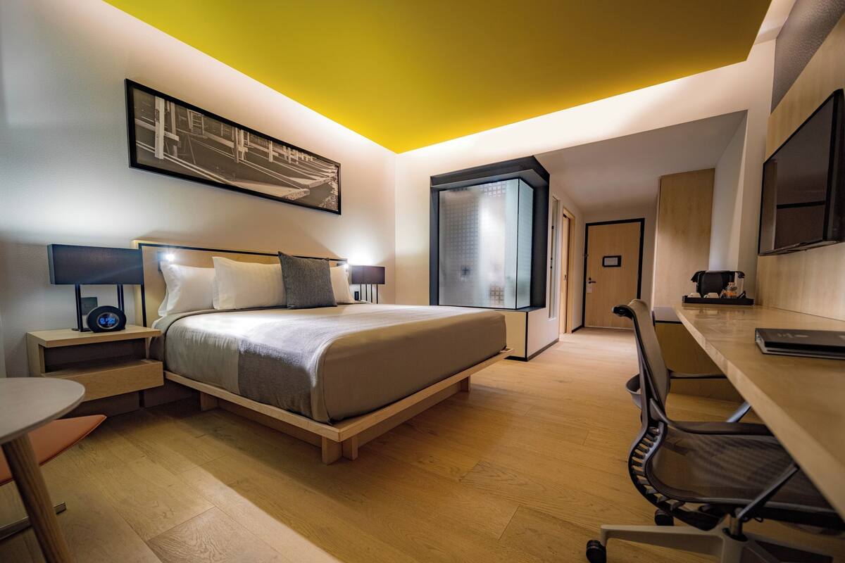 Une chambre d’hôtel moderne équipée d’un lit, d’une table de chevet, d’une lampe, d’un bureau, d’une chaise et d’une grande fenêtre.