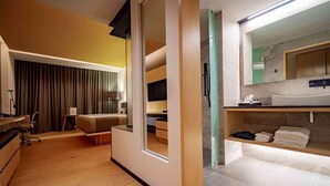 Moderni hotellihuone, jossa on liukuovi, joka johtaa parvekkeelle, erillinen oleskelualue, jossa on sohva ja työpöytä, sekä kylpyhuone, jossa on suuri peili ja pesuallas.