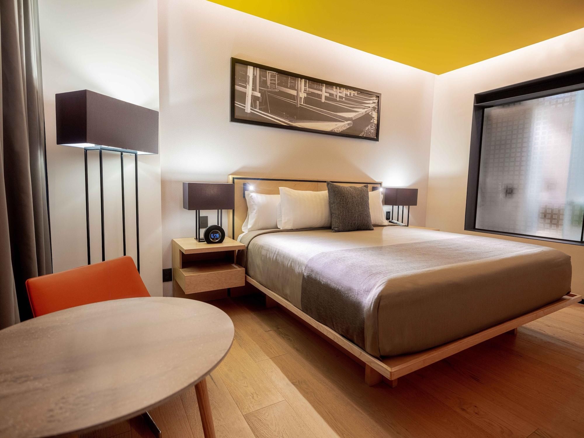 A hotel room with a bed, bedside tables, a nightstand, a chair, a round table, a window, and a wall-mounted artwork.