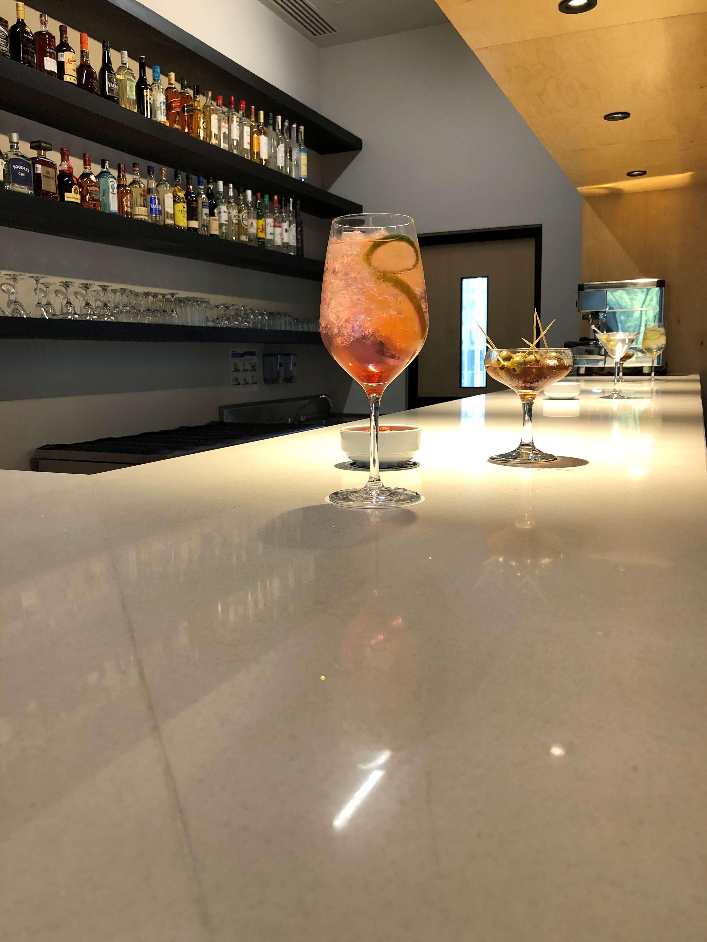 A bar area with a polished countertop, a variety of liquor bottles on shelves, and two glasses of cocktails.