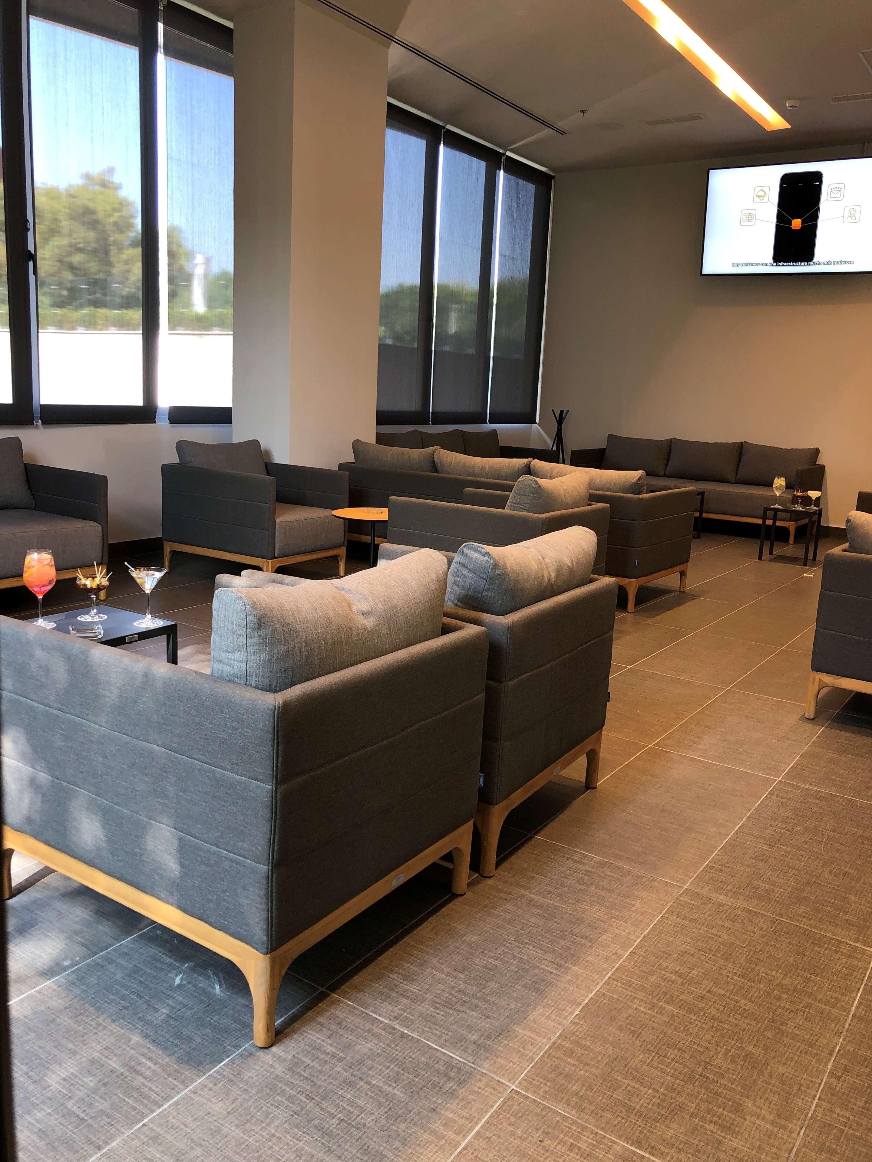 A modern lounge area with multiple sofas and armchairs, a flat-screen TV mounted on the wall, and large windows providing natural light.