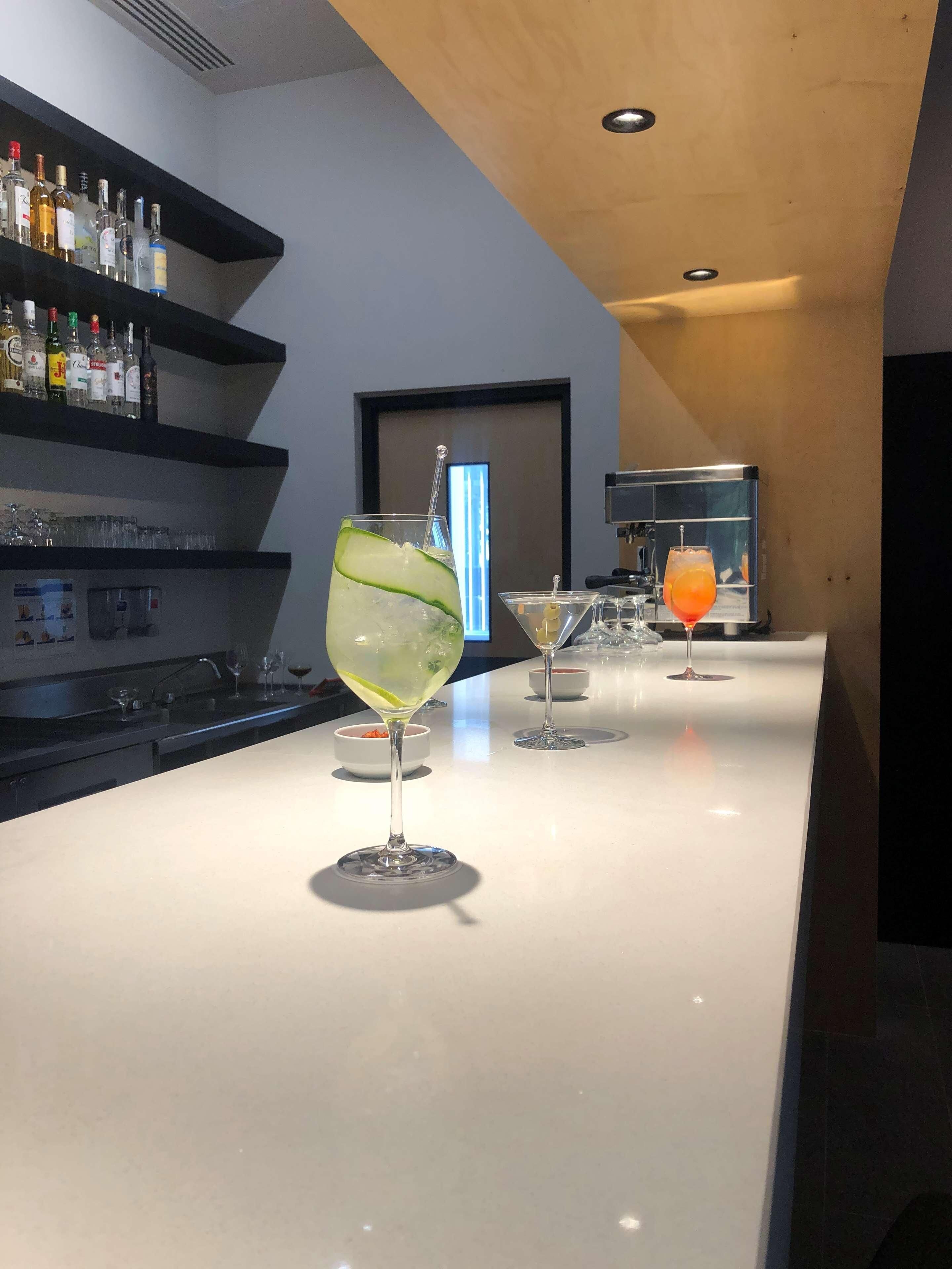 Zona de bar con mostrador blanco, vasos y botellas. El techo es de madera y hay un estante negro con botellas.