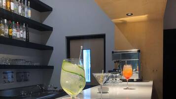 Zona de bar con mostrador blanco, vasos y botellas. El techo es de madera y hay un estante negro con botellas.