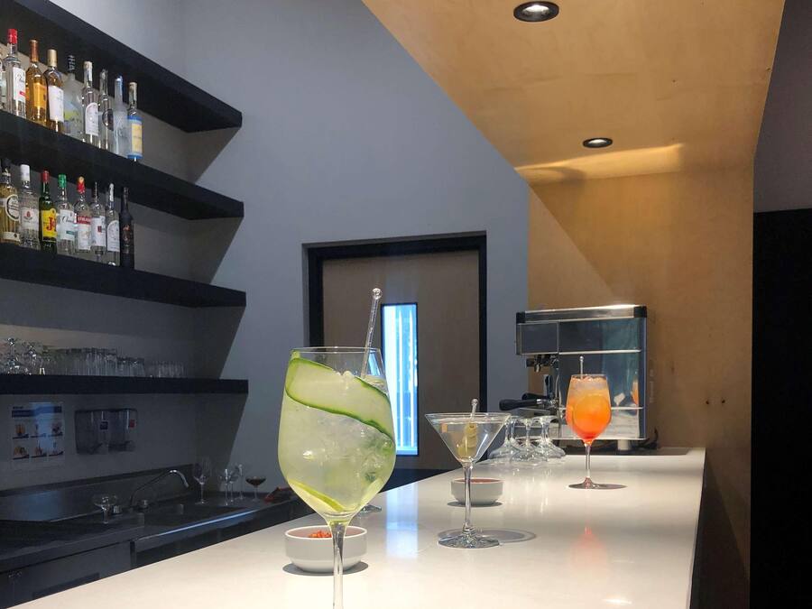 Zona de bar con mostrador blanco, vasos y botellas. El techo es de madera y hay un estante negro con botellas.