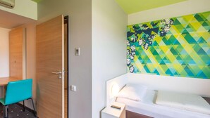A hotel room with a wooden door, a teal chair, a bed with a white headboard, a nightstand, and a colorful wall mural. - B&B Hotel Erlangen (Erlangen)