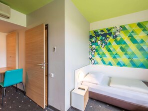 A hotel room with a wooden door, a teal chair, a bed with a white headboard, a nightstand, and a colorful wall mural. - B&B Hotel Erlangen (Erlangen)