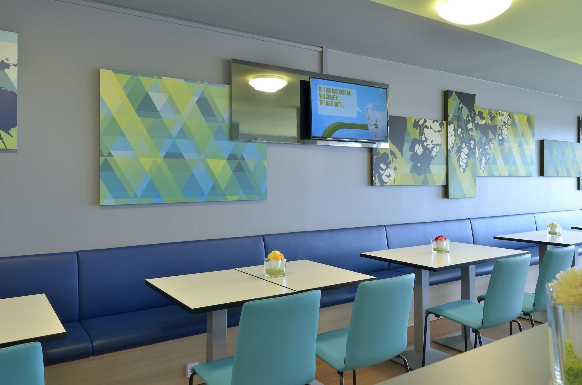 A modern restaurant interior with blue and green geometric wall art, a flat-screen TV displaying a welcome message, and a seating area with light blue chairs and white tables.