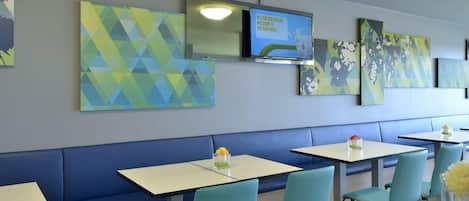 Un interior de restaurante moderno con arte mural geométrico en tonos azules y verdes, un televisor de pantalla plana mostrando un mensaje de bienvenida, y una zona de estar con sillas azul claro y mesas blancas.