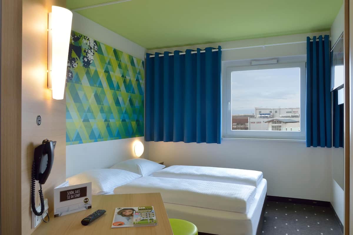 A hotel room with two beds, a desk with a phone and a remote control, a wall clock, and a window with curtains.