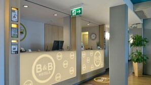 A hotel reception area with a reception desk, a clock, and a green exit sign. - B&B Hotel Erlangen (Erlangen)