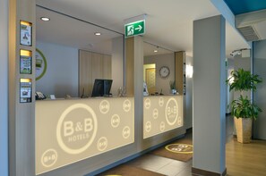 A hotel reception area with a reception desk, a clock, and a green exit sign. - B&B Hotel Erlangen (Erlangen)
