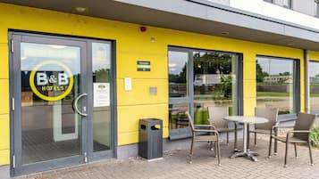 A hotel entrance with a yellow facade, a glass door, and a small outdoor seating area with wicker chairs and a table.