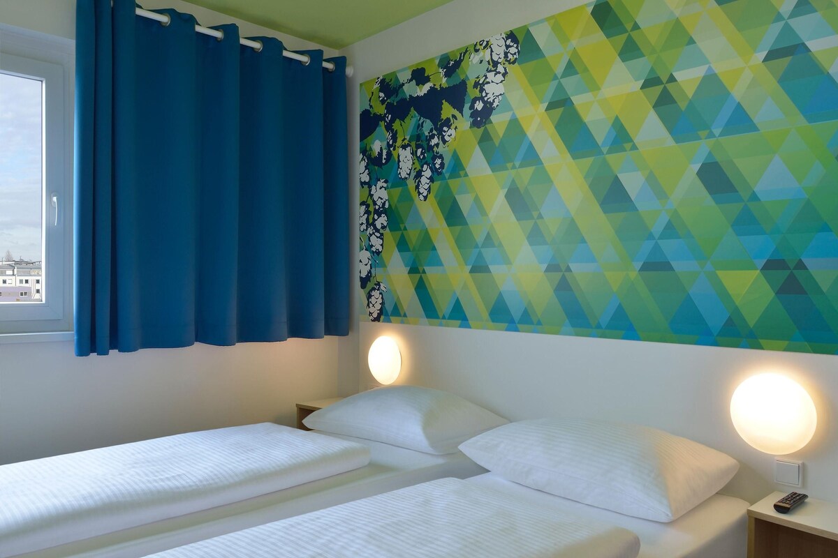 A hotel room with two beds, a window with blue curtains, a wall with a geometric pattern, and two bedside lamps.
