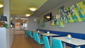 A modern restaurant with teal chairs and white tables, featuring geometric wall art and a visible kitchen area. - B&B Hotel Erlangen (Erlangen)
