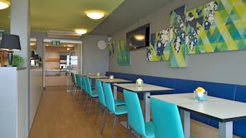 A modern restaurant with teal chairs and white tables, featuring geometric wall art and a visible kitchen area.