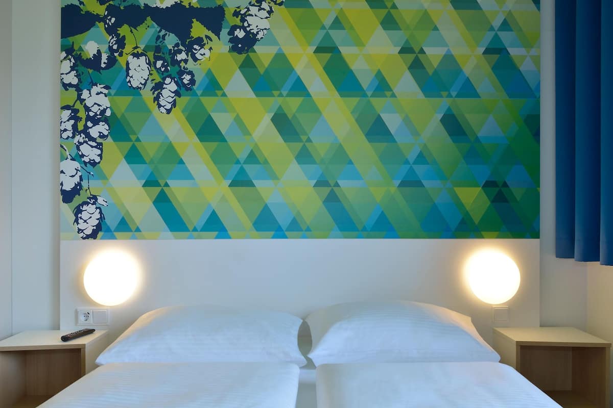 A hotel room with a bed, two bedside tables, a wall-mounted lamp, and a geometric-patterned wall decoration.