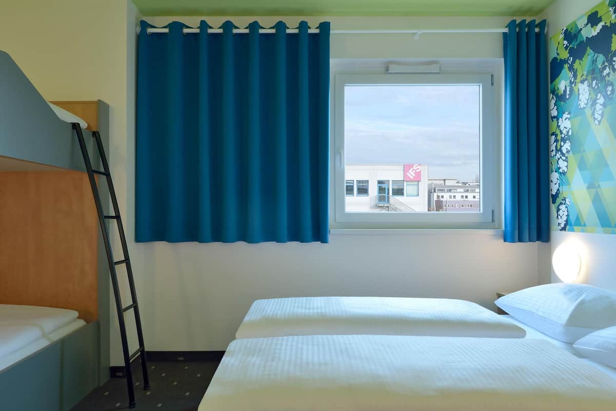 A hotel room with a bed, a window with blue curtains, a ladder, and a wall-mounted light.