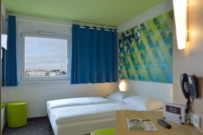 A hotel room with a bed, a window with curtains, a wall-mounted phone, and a magazine on the table.
