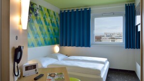 A hotel room with two beds, a desk with a phone and a remote control, a wall clock, and a window with curtains. - B&B Hotel Erlangen (Erlangen)