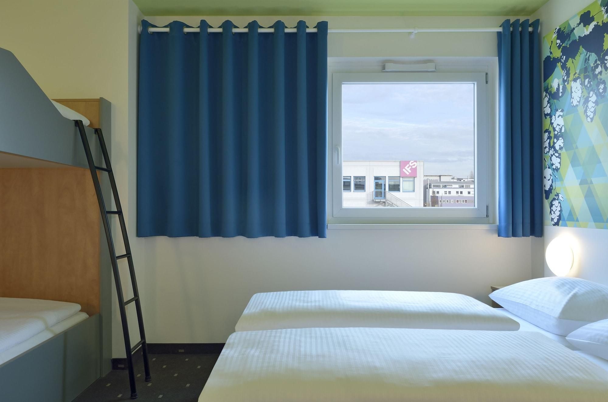 A hotel room with a bed, a window with curtains, a bedside lamp, and a view of a building outside.