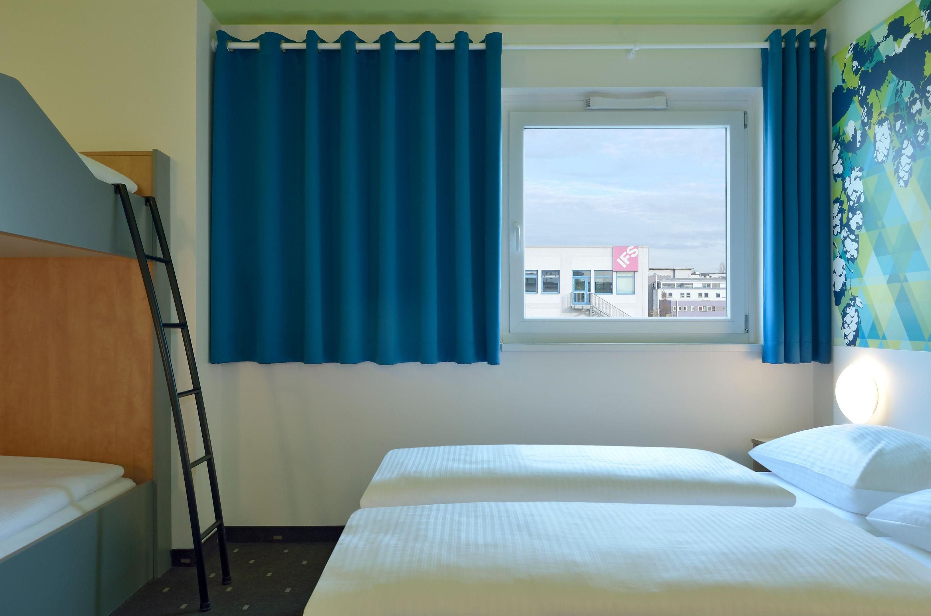 A hotel room with a bed, a window with blue curtains, a ladder, and a wall-mounted light.