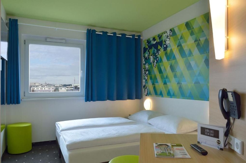 Una habitación de hotel con cama, ventana con cortinas azules, un taburete verde, teléfono montado en la pared y un colorido cuadro abstracto geométrico.