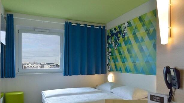A hotel room with a bed, a window with blue curtains, a green stool, a wall-mounted phone, and a colorful geometric wall art. - B&B Hotel Erlangen (Erlangen)