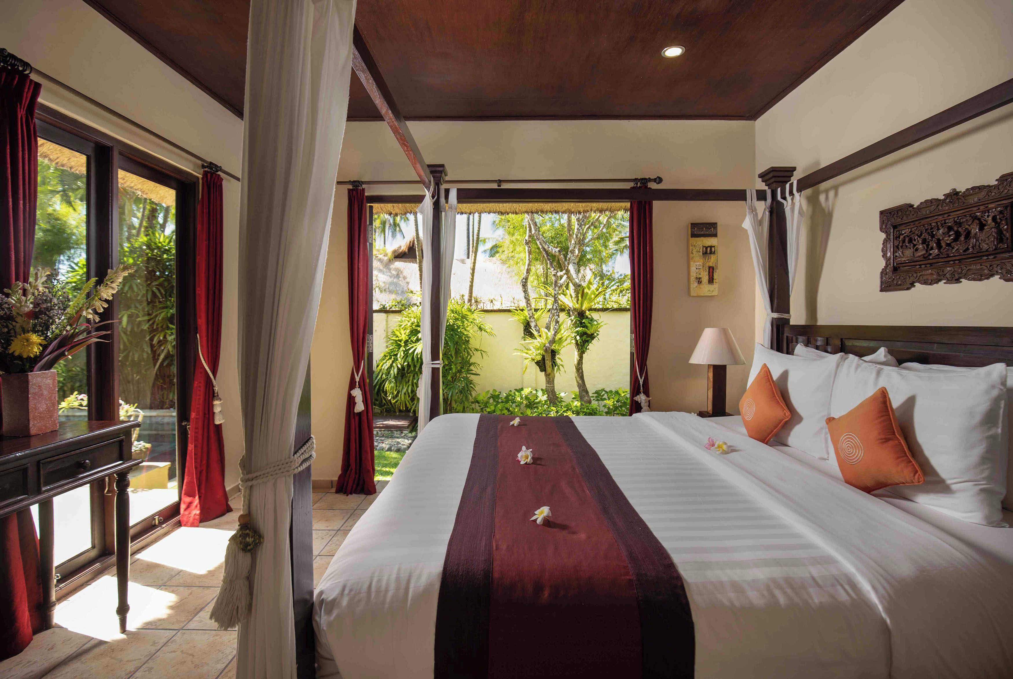 A hotel room with a large bed, a nightstand, a window with curtains, and a view of greenery outside.