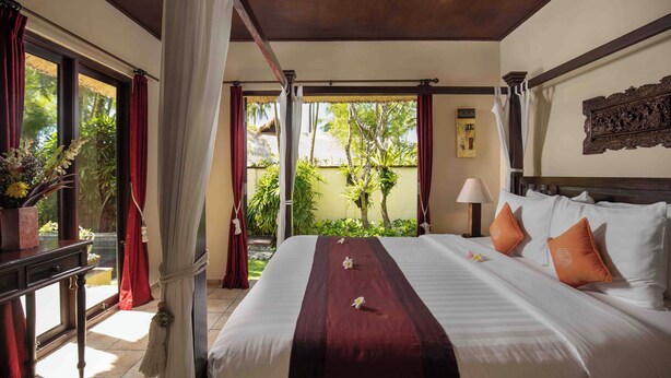 A hotel room with a large bed, a nightstand, a window with curtains, and a view of greenery outside. - Alam Anda Ocean Front Resort & Spa (Tejakula)