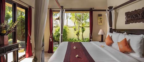 A hotel room with a large bed, a nightstand, a window with curtains, and a view of greenery outside.