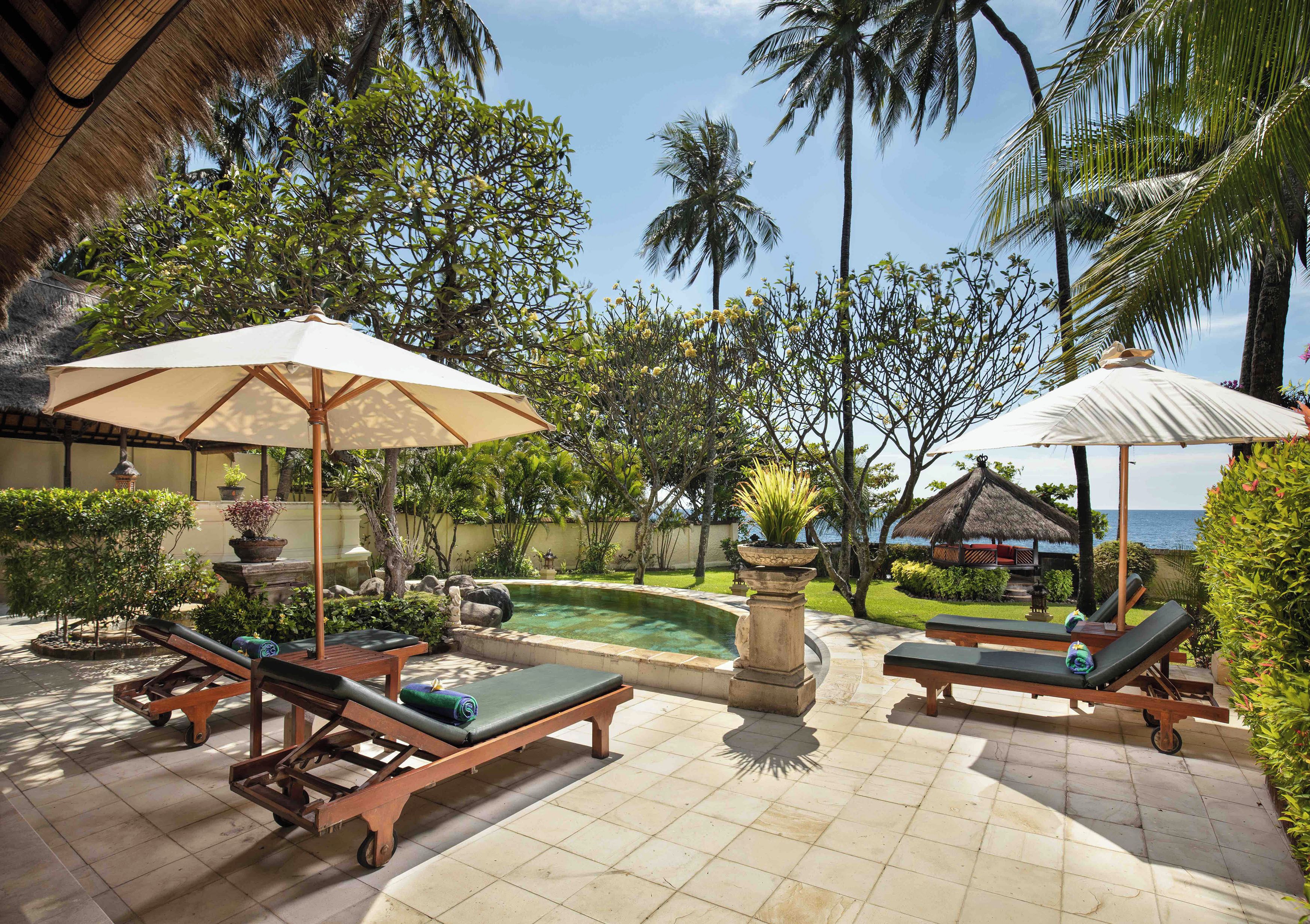 Zona de piscina con sillones reclinables y sombrillas, rodeada de vegetación tropical y vista al océano.