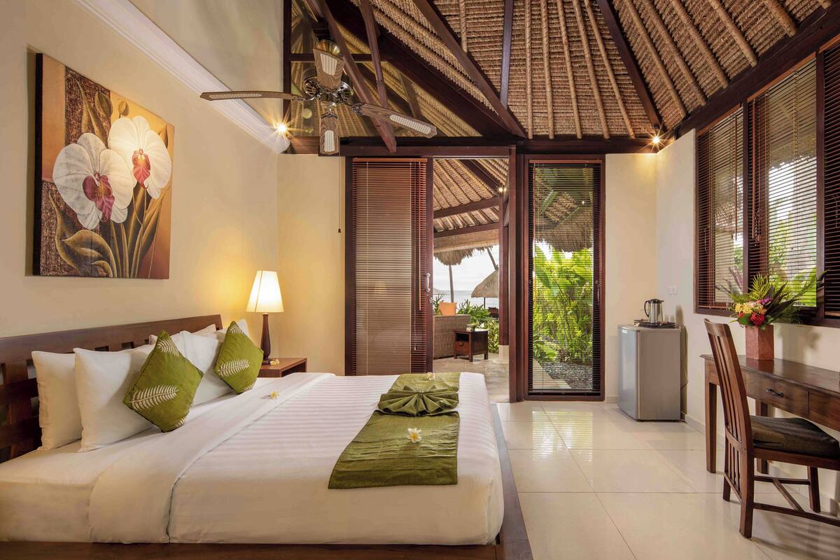 A hotel room with a bed, a desk, a chair, a ceiling fan, a painting, and a view of the outdoors.