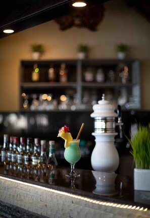 Un’area bar con un cocktail sul bancone, scaffali con bottiglie e un vaso bianco decorativo.