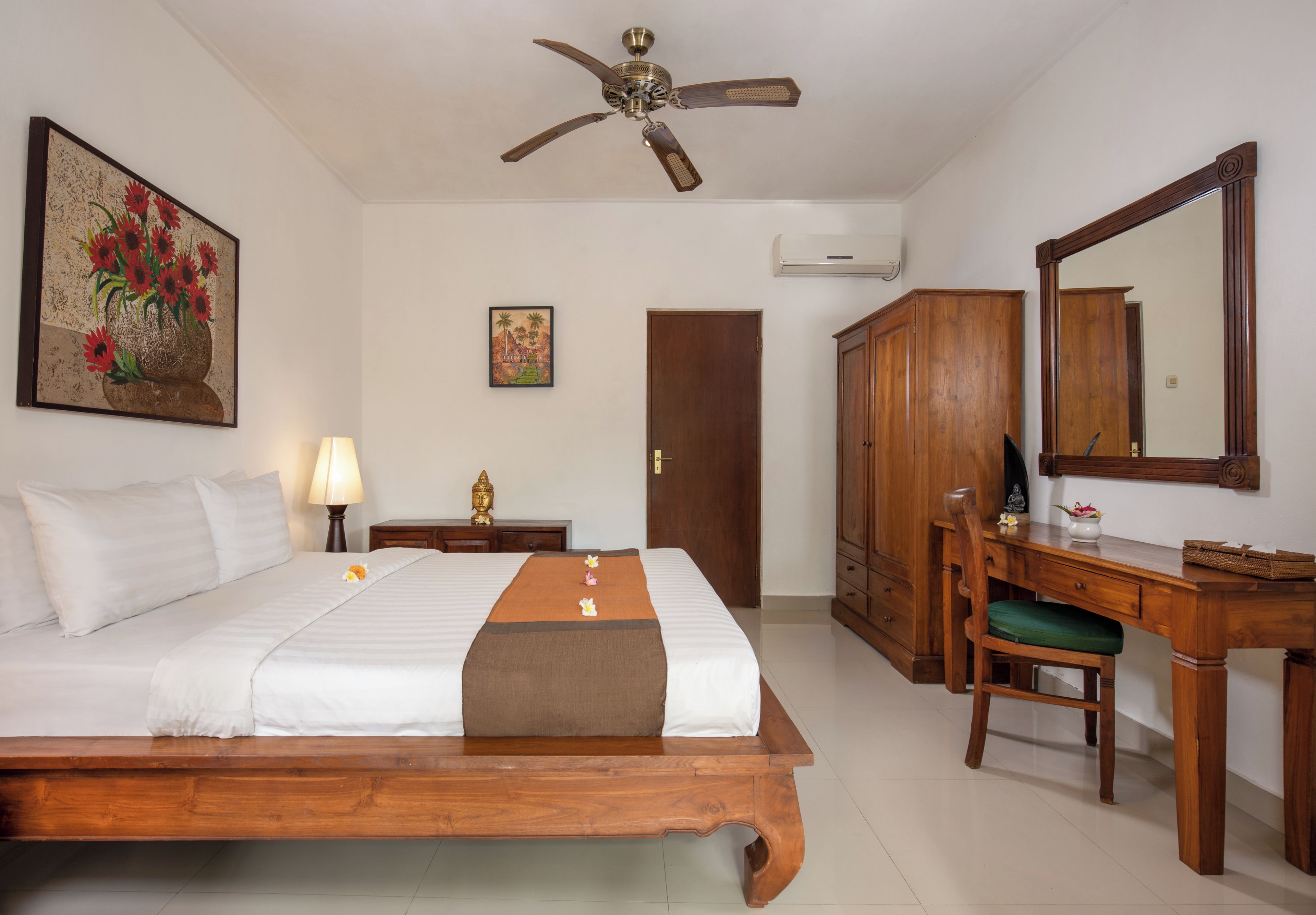 A hotel room with a wooden bed, a desk with a chair, a ceiling fan, a large mirror, and artwork on the walls.