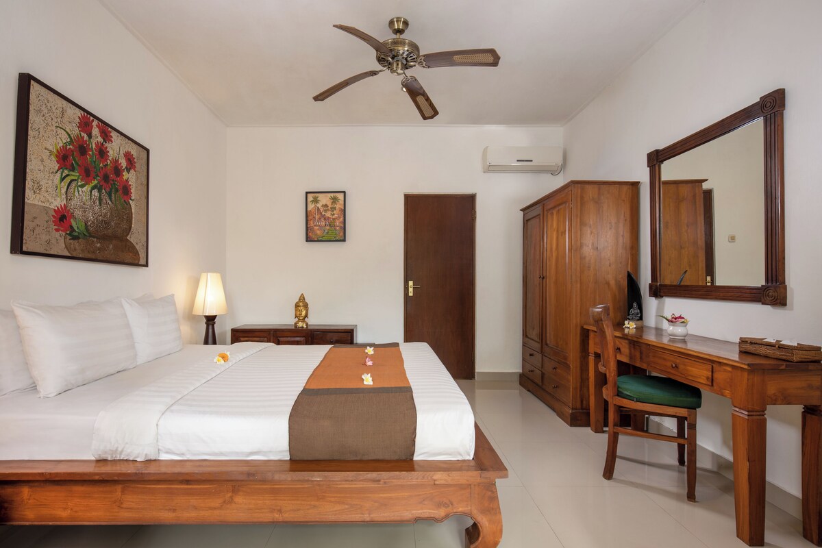 A hotel room with a wooden bed, a desk with a chair, a ceiling fan, a large mirror, and artwork on the walls.