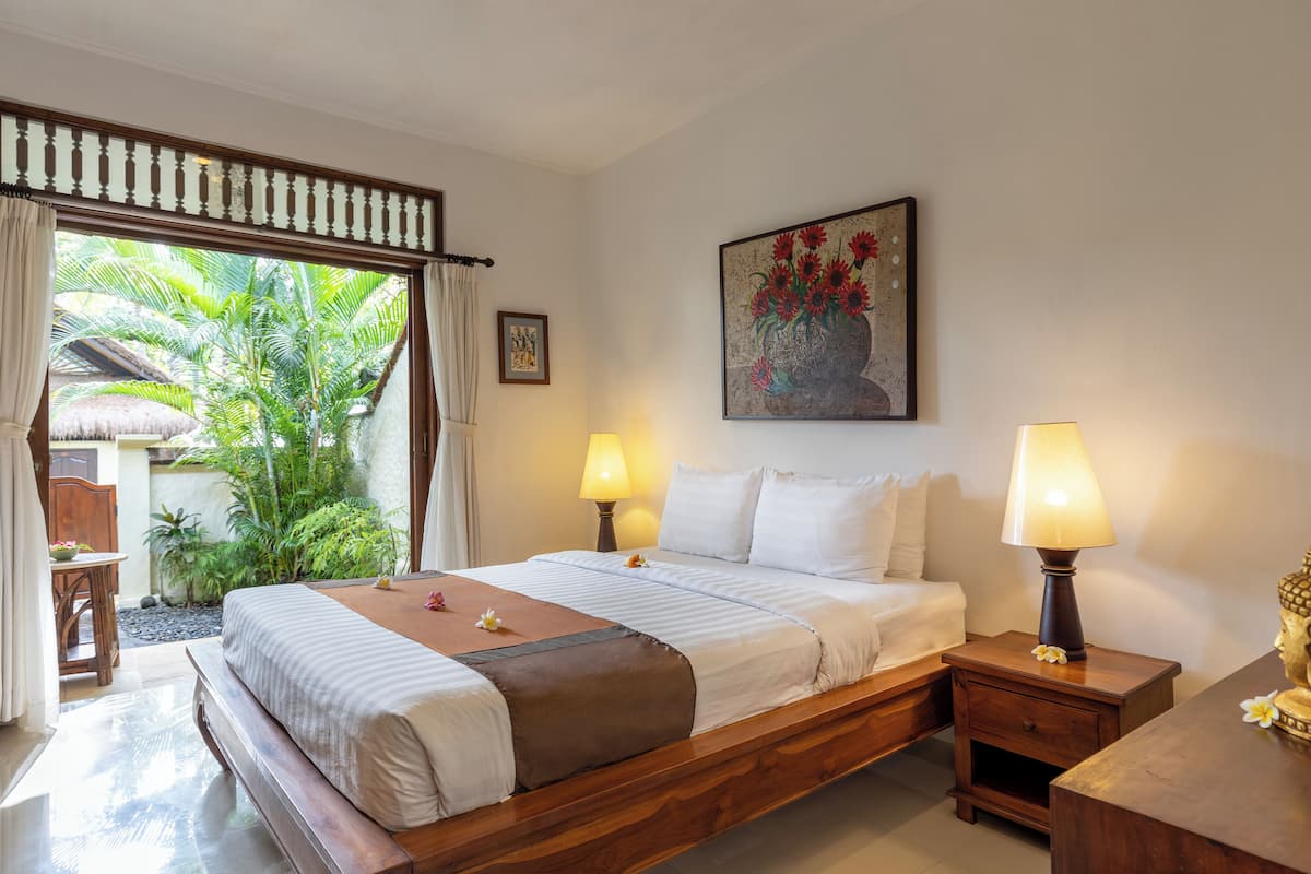 A bedroom with a bed, bedside tables, a nightstand, a painting, and a view of greenery outside.