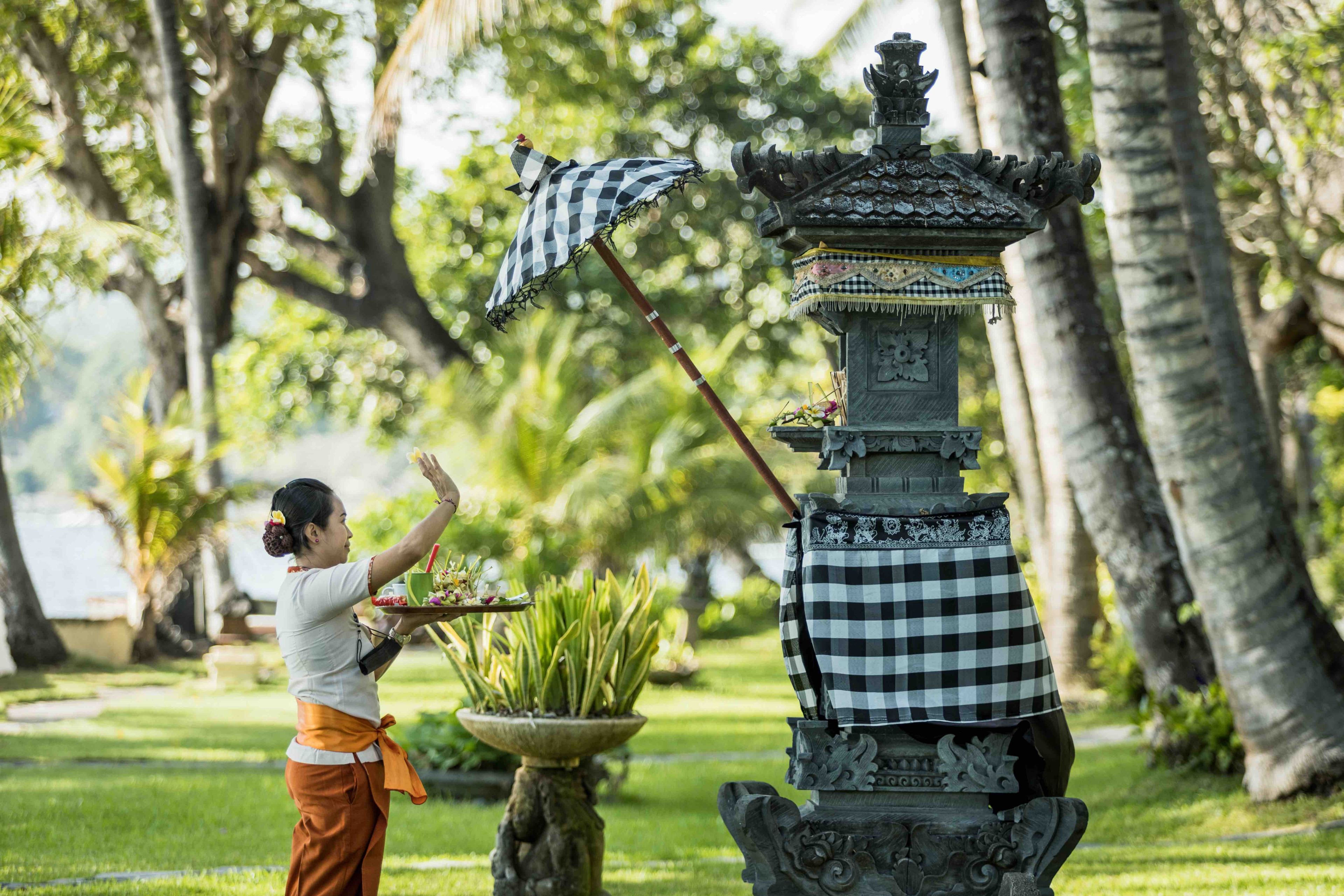 Uma pessoa realizando um ritual com um guarda-chuva xadrez e oferendas em uma estrutura de pedra tradicional em um jardim exuberante.
